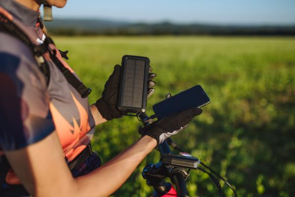 Cyclist charging her smartphone using a solar phone charger. Female cyclist standing in front of solar panels at a solar farm during a summer bike tour in nature. Concept of sustainable energy future.