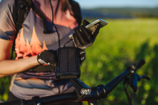 Cyclist charging her smartphone using a solar phone charger. Female cyclist standing in front of solar panels at a solar farm during a summer bike tour in nature. Concept of sustainable energy future.