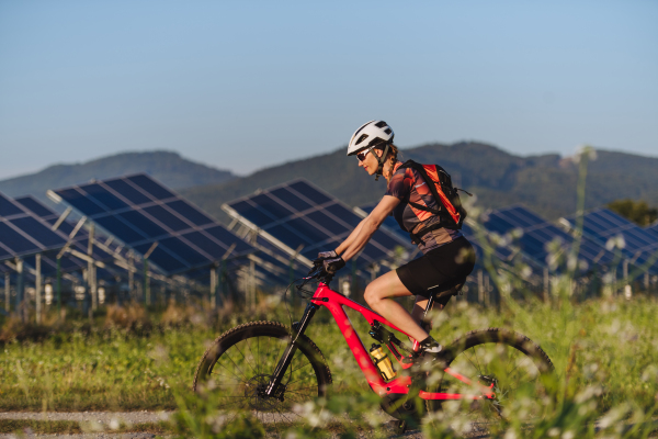 Side view of a beautiful cyclist riding in front of solar panels at a solar farm during a summer bike tour in nature. A solar farm as solution for more sustainable energy future.