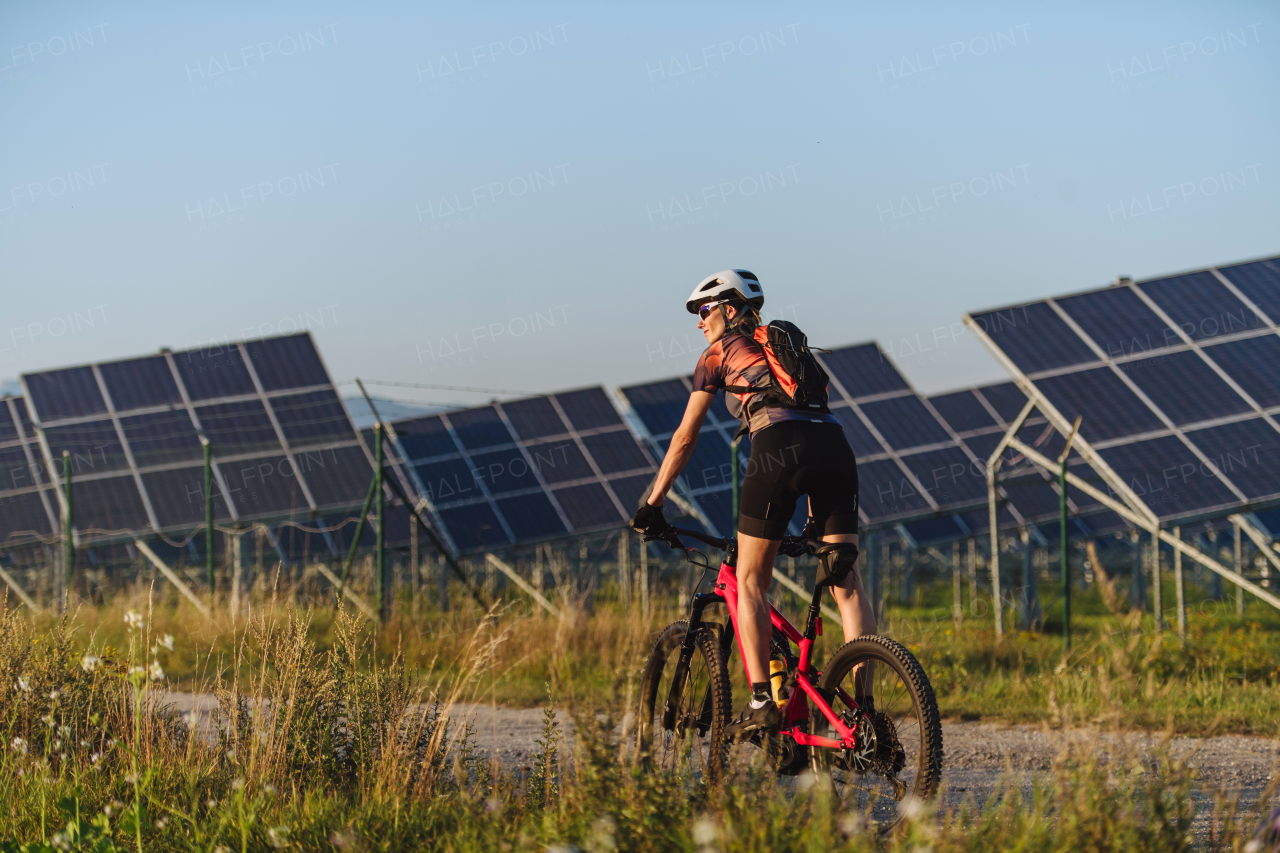 Rear view of a beautiful cyclist riding in front of solar panels at a solar farm during a summer bike tour in nature. A solar farm as solution for more sustainable energy future.