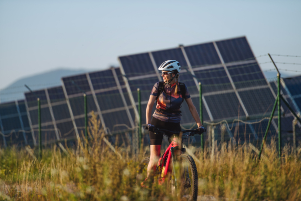 Beautiful cyclist riding in front of solar panels at a solar farm during a summer bike tour in nature. A solar farm as solution for more sustainable energy future.