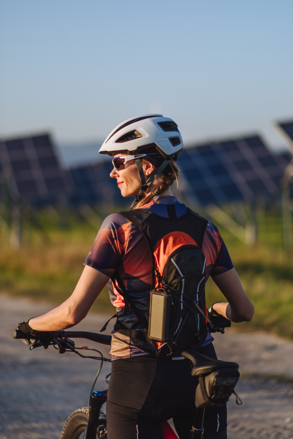 Rear view of a beautiful cyclist standing in front of solar panels at a solar farm during a summer bike tour in nature. A solar farm as solution for more sustainable energy future.