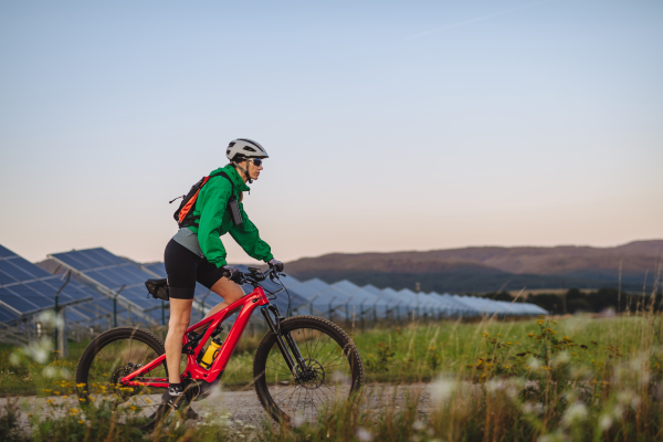 Side view of a beautiful cyclist riding in front of solar panels at a solar farm during a summer bike tour in nature. A solar farm as solution for more sustainable energy future.