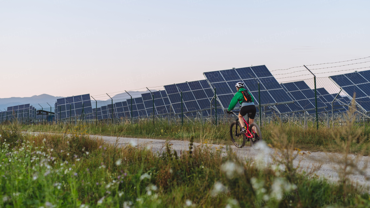 Rear view of a female cyclist riding in front of solar panels at a solar farm during a summer bike tour in nature. A solar farm as solution for more sustainable energy future. Banner with copy space.