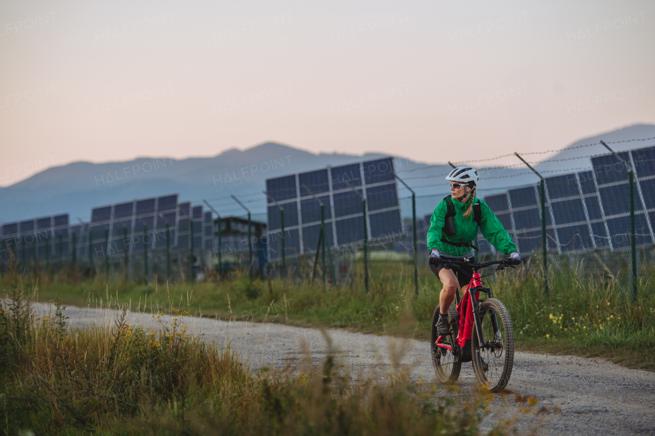 Female cyclist riding in front of solar panels at a solar farm during a summer bike tour in nature. A solar farm as solution for more sustainable energy future. Banner with copy space.