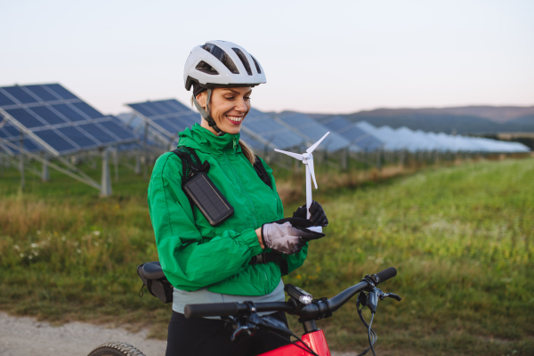 Portrait of a beautiful cyclist standing in front of solar panels at a solar farm during a summer bike tour in nature. Cyclist holding model of wind turbine. Alternative and sustainable energy sources.