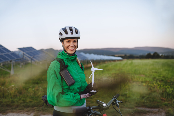 Portrait of a beautiful cyclist standing in front of solar panels at a solar farm during a summer bike tour in nature. Cyclist holding model of wind turbine. Alternative and sustainable energy sources.