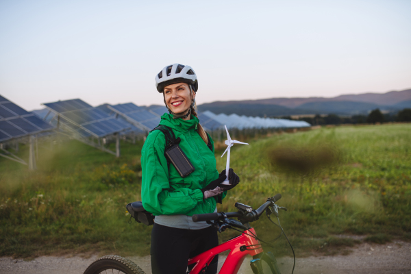 Portrait of a beautiful cyclist standing in front of solar panels at a solar farm during a summer bike tour in nature. Cyclist holding model of wind turbine. Alternative and sustainable energy sources.