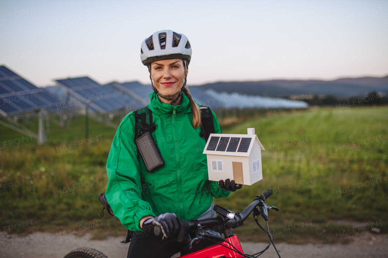 Portrait of a beautiful cyclist standing in front of solar panels at a solar farm during a summer bike tour in nature. Cyclist holding model of house with photovoltaic panels on roof. Solar energy sustainable energy future.