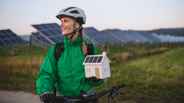 Portrait of a beautiful cyclist standing in front of solar panels at a solar farm during a summer bike tour in nature. Cyclist holding model of house with photovoltaic panels on roof. Solar energy sustainable energy future.