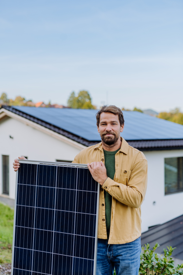 Mature man holding a solar panel near his house with solar panels on the roof. Alternative energy, saving resources and sustainable lifestyle concept.