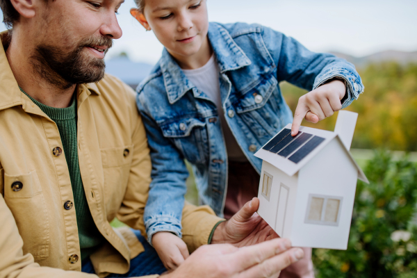 Little girl with her father holding paper model of house with the solar panels, explaining how it works.Alternative energy, saving resources and sustainable lifestyle concept.