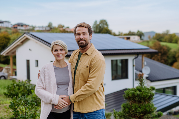 Happy man with his pregnant wife standing in front of their house with photovoltaics solar panels.