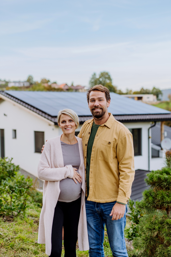 Happy man with his pregnant wife standing in front of their house with photovoltaics solar panels.