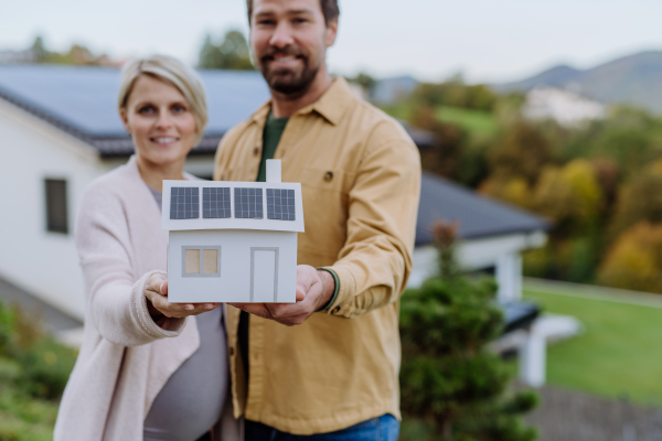 Close-up of happy couple holding paper model of house with solar panels. Alternative energy, saving resources and sustainable lifestyle concept.