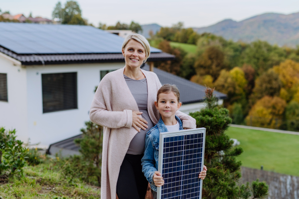 Mother with her little daughter near the house with solar panels. Alternative energy, saving resources and sustainable lifestyle concept.