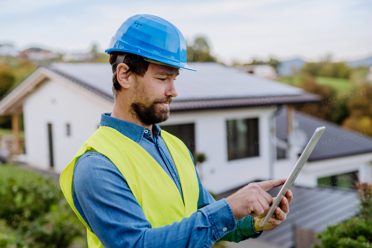 Smiling handyman solar installer standying in front of family house with solar system on the roof and checking a digital dablet.