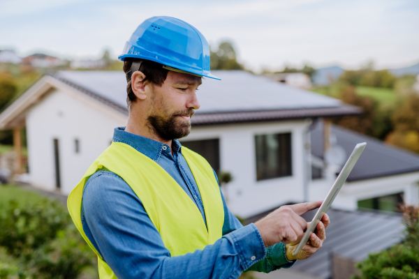 Smiling handyman solar installer standying in front of family house with solar system on the roof and checking a digital dablet.