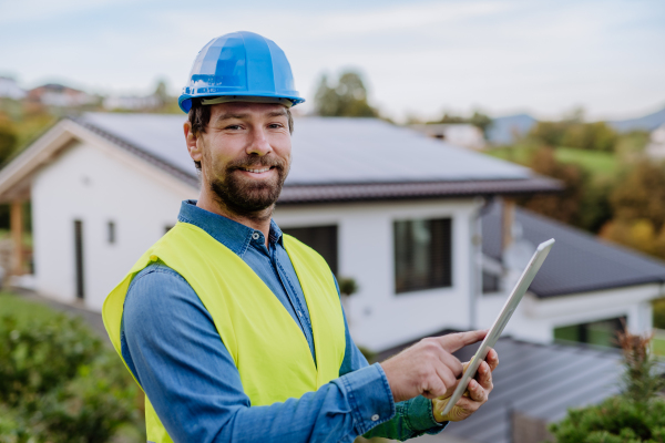 Smiling handyman solar installer standying in front of family house with solar system on the roof and checking a digital dablet.