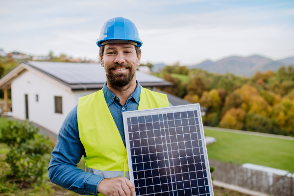 A smiling handyman solar installer carrying solar module while installing solar panel system on house.