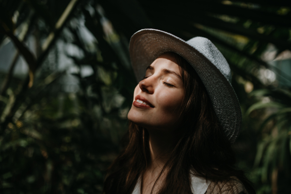 Portrait of a young woman with hat and closed eyes in botanical garden. Beautiful woman botanist in jungle, side view.