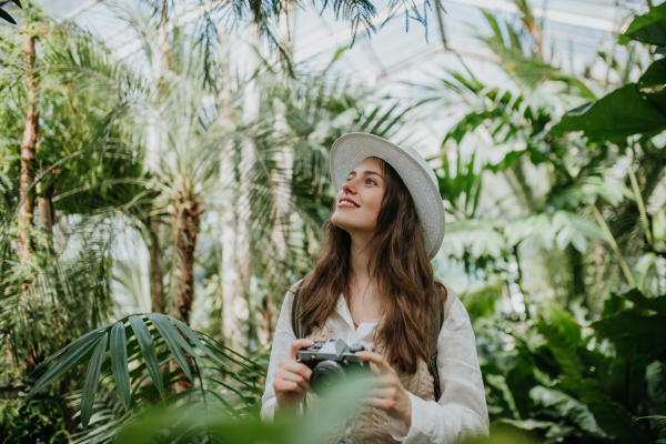 Portrait of a young woman photographer with hat and backpack in botanical garden. Botanist in greenhouse taking pictures.