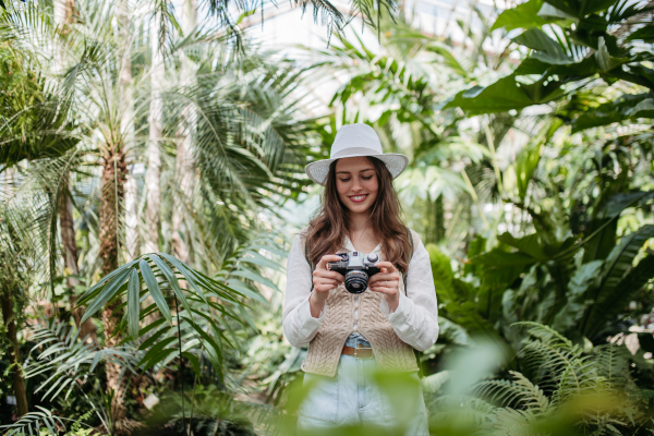 Portrait of a young woman photographer with hat and backpack in botanical garden. Botanist in greenhouse taking pictures.