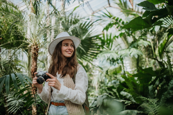 Portrait of a young woman photographer with hat and backpack in botanical garden. Botanist in greenhouse taking pictures.