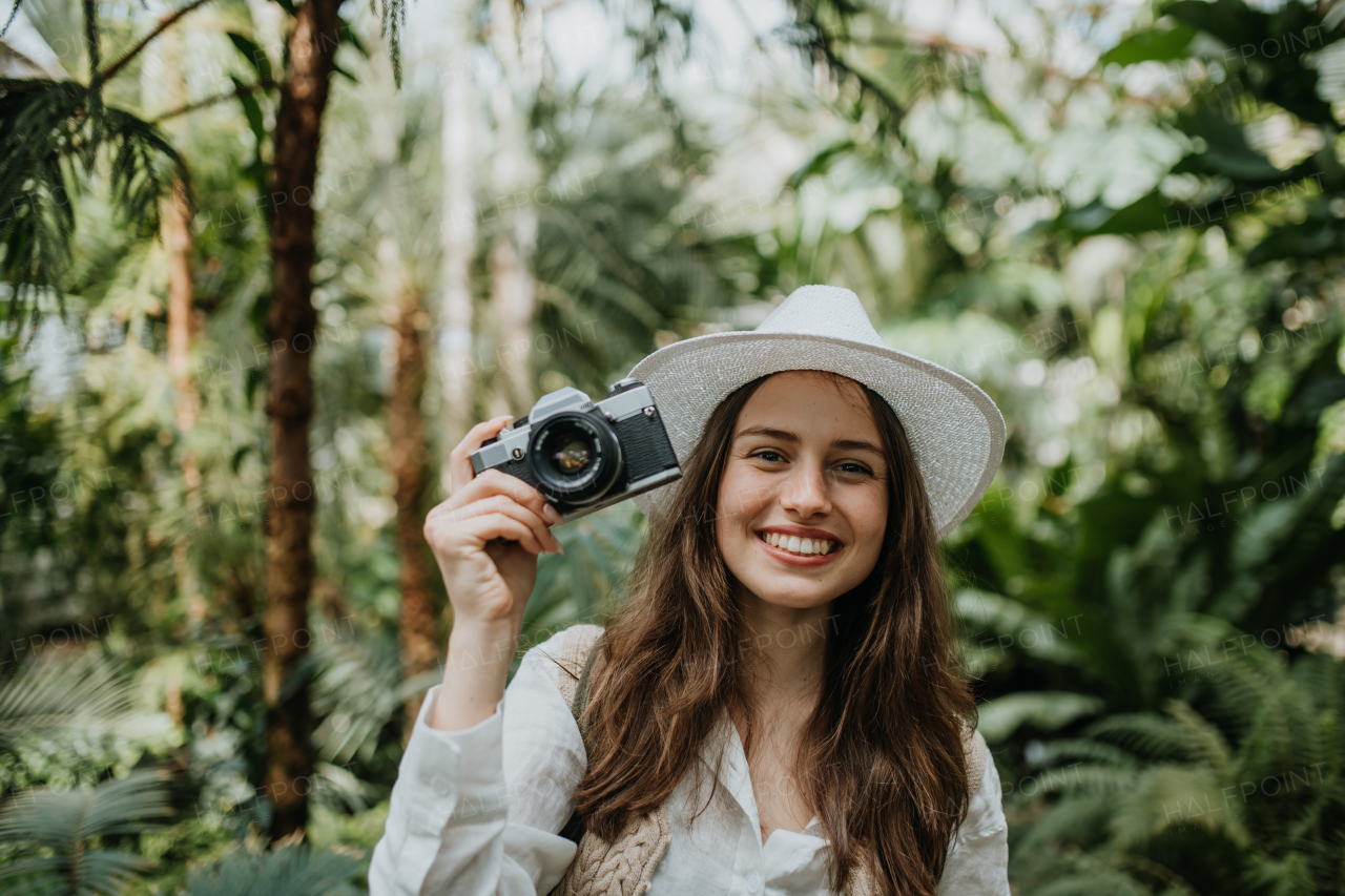 Portrait of a young woman photographer with hat and backpack in botanical garden. Botanist in greenhouse taking pictures.