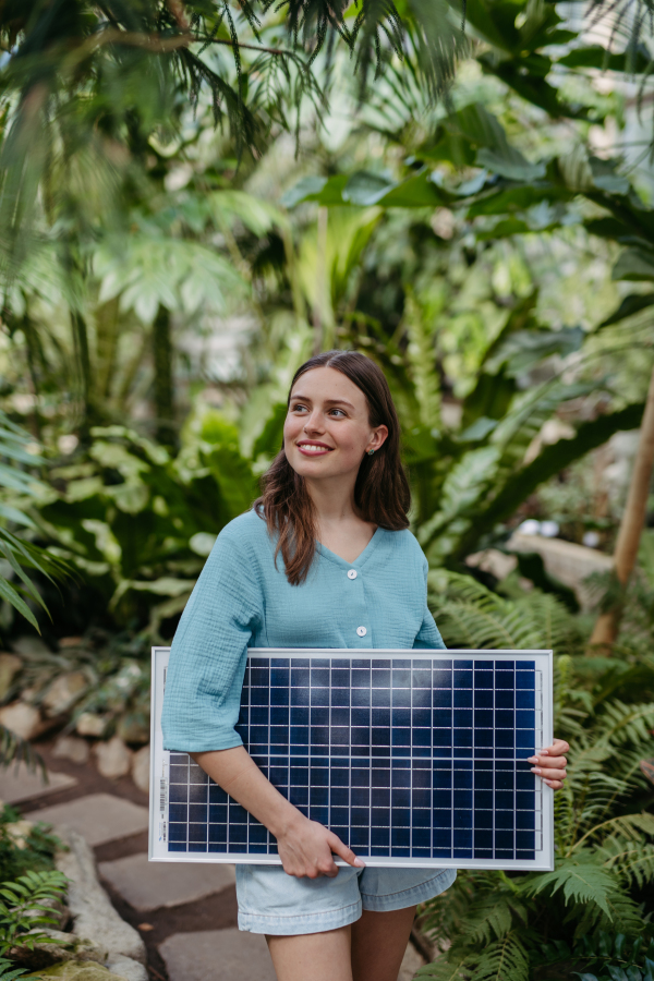 Young woman in greenhouse holding model of house with solar panels, concept of renewable energy and protection of nature.