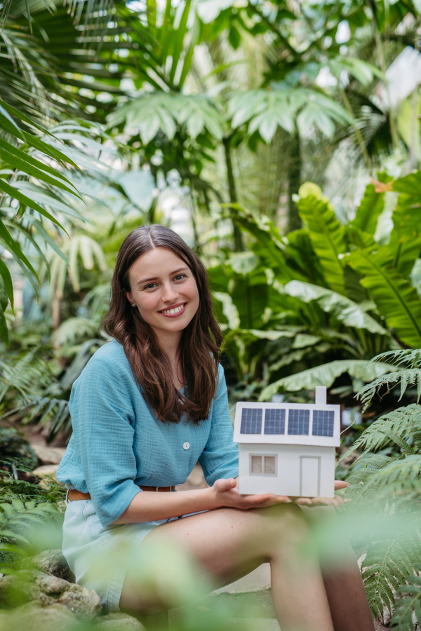 Young woman in jungle holding model of house with solar panels, concept of renewable energy and protection of nature.