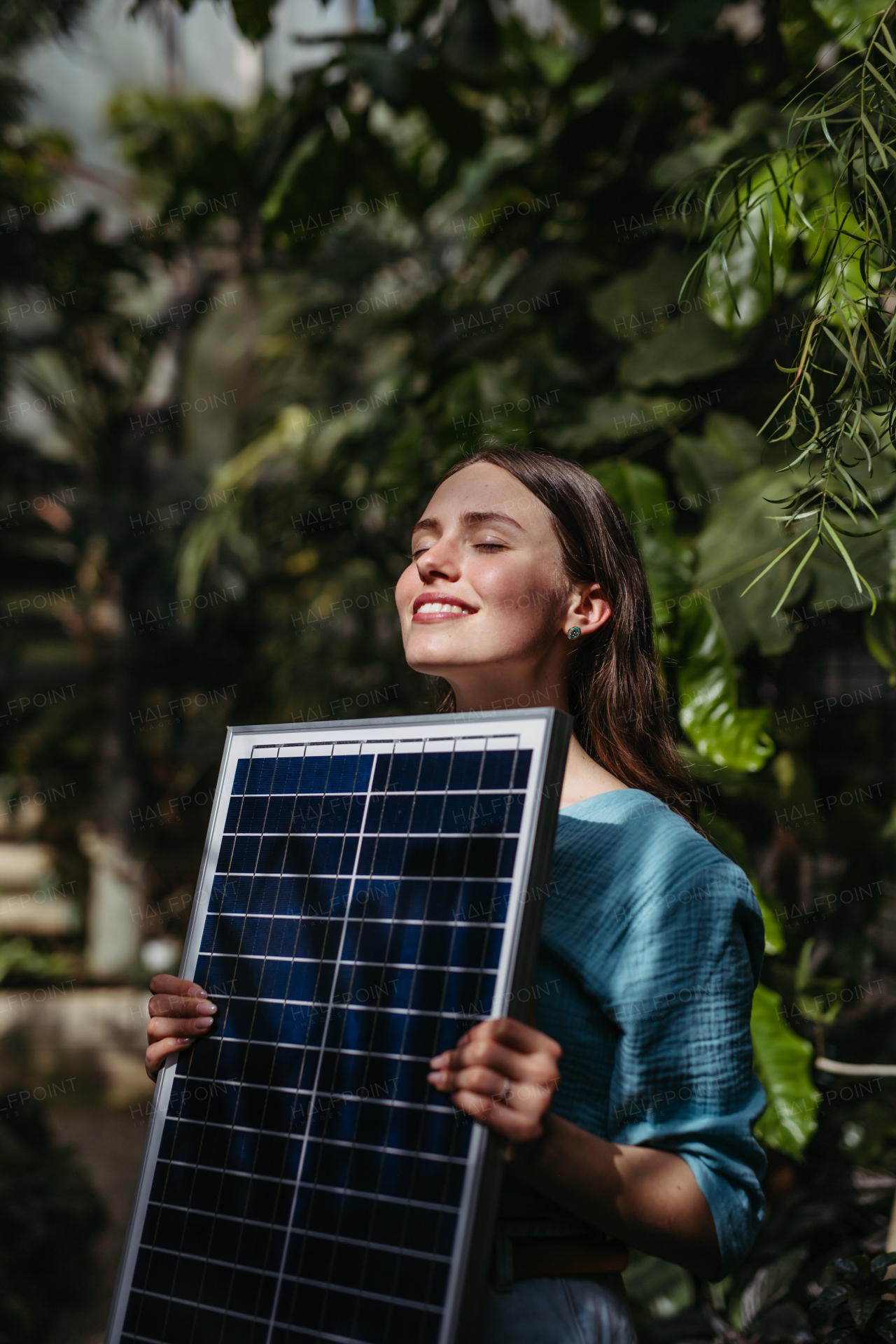 Young woman in greenhouse holding model of house with solar panels, concept of renewable energy and protection of nature.