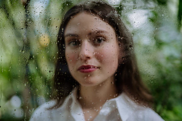 Portrait of a young woman in misty greenhouse through foggy glass with water droplets.