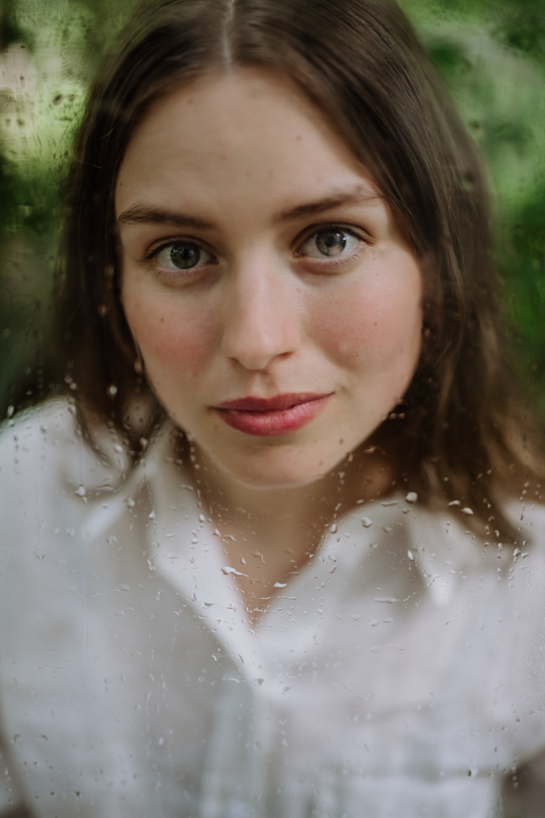 Portrait of a young woman in misty greenhouse through foggy glass with water droplets.