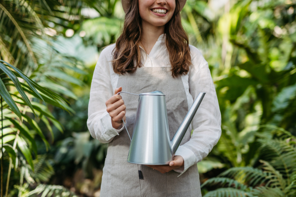 Portrait of beautiful female gardener taking care of plants in botanical garden. Botanist, horticulturist watering plants with watering can.