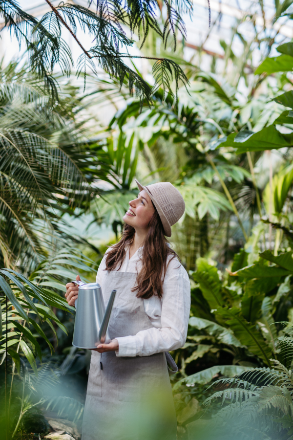 Portrait of beautiful female gardener taking care of plants in botanical garden. Botanist, horticulturist watering plants with watering can.