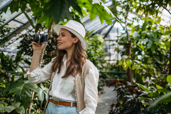 Portrait of a young woman photographer with hat and backpack in botanical garden. Botanist in greenhouse taking pictures.