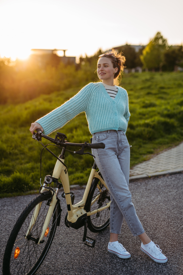 Young woman standing by electro bicycle, during sunset, sunrise. Concept of commuting and ecologic traveling.