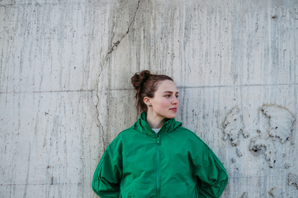 Waist up portrait of young city woman in green windbreaker jacket leaning against a concrete wall during sunset. Copy space image.