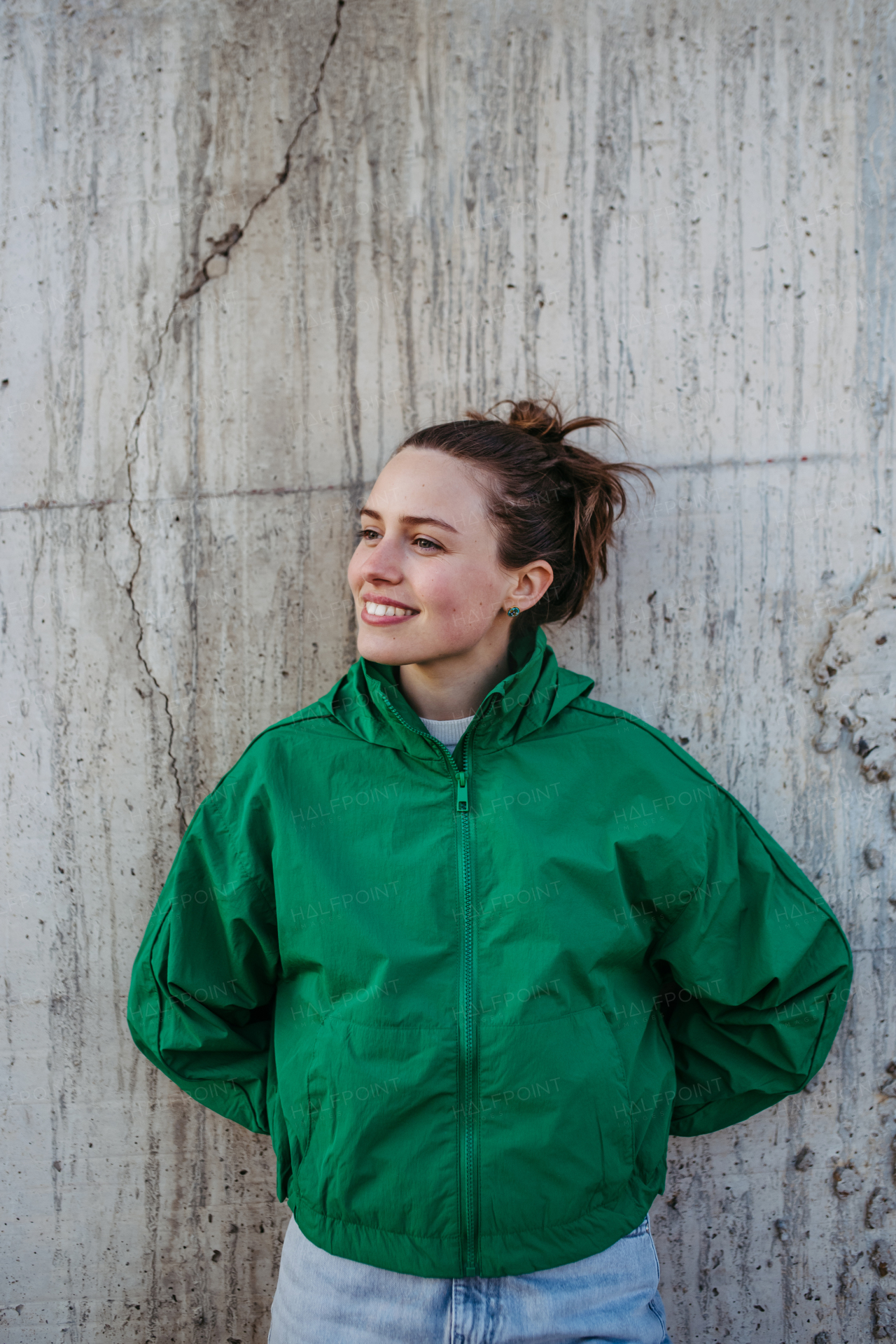 Waist up portrait of young city woman in green windbreaker jacket leaning against a concrete wall during sunset. Copy space image.