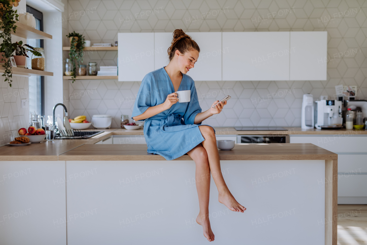 Young woman with smartphone enjoying cup of coffee at morning, in a kitchen.