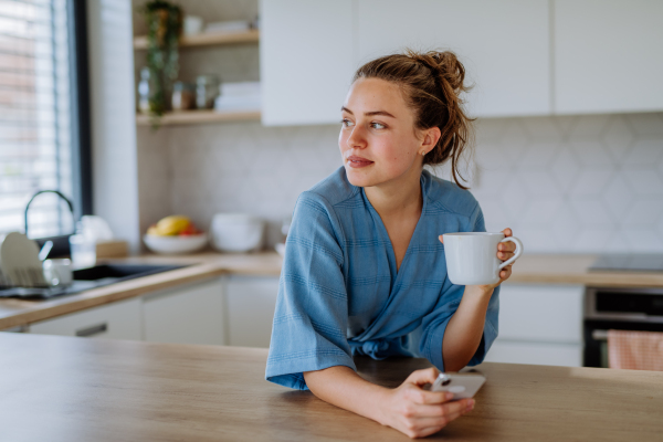 Young woman with smartphone enjoying cup of coffee at morning, in a kitchen.