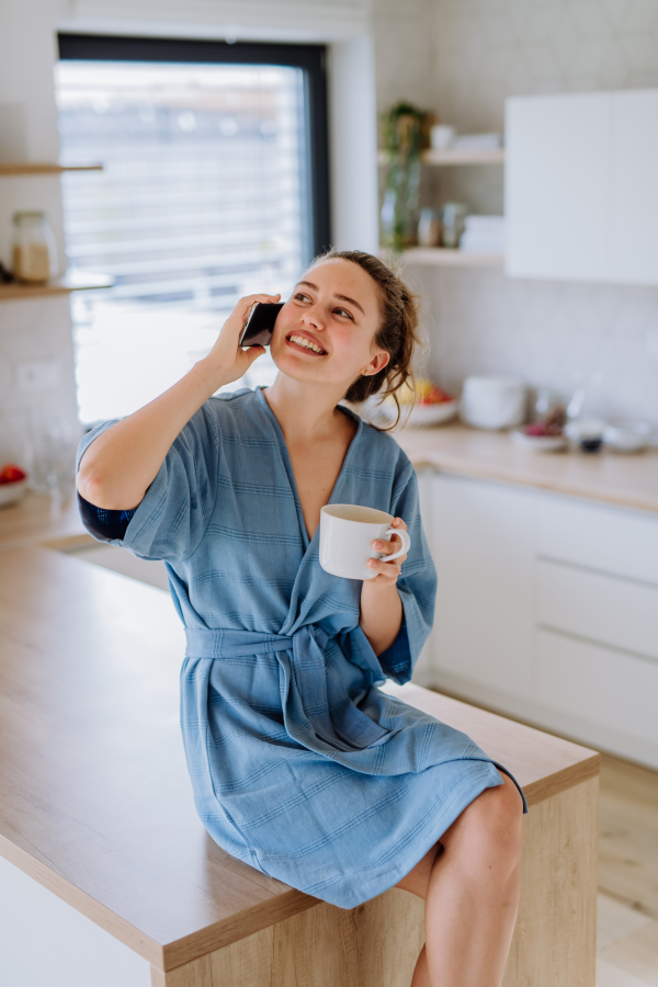 Young woman calling and enjoying cup of coffee at morning, in a kitchen.