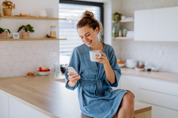 Young woman with smartphone enjoying cup of coffee at morning, in a kitchen.