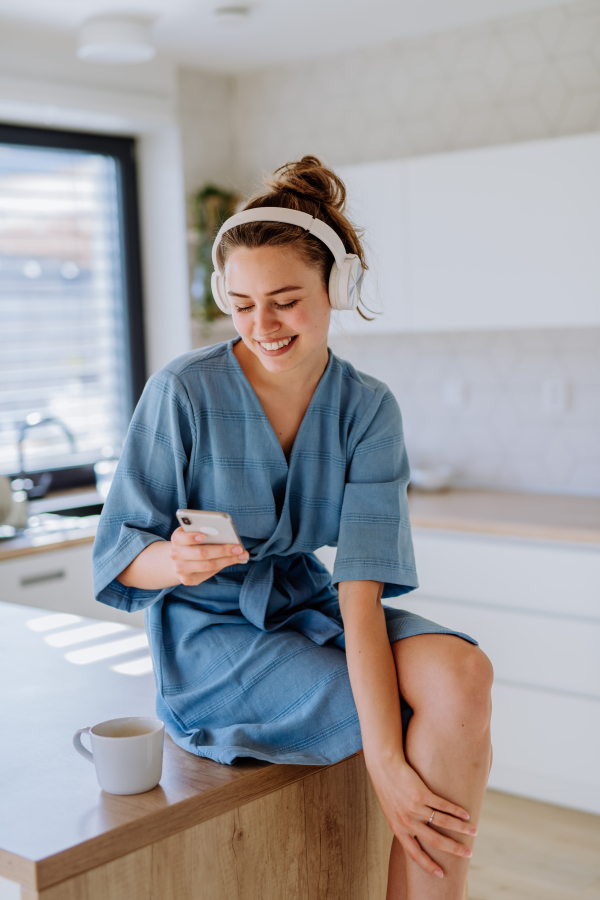 Young woman enjoying cup of coffee,scrolling smartphone and listening music at morning, in a kitchen.