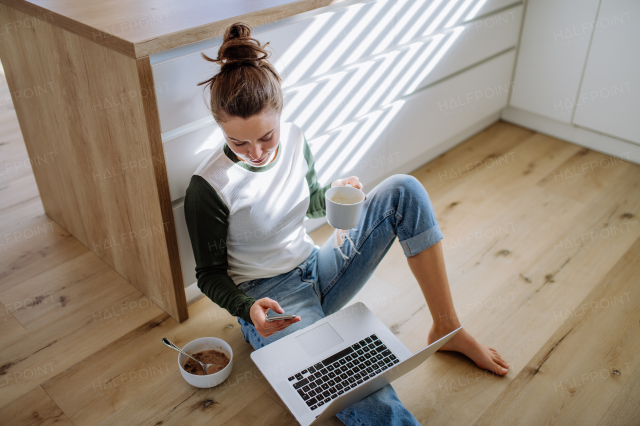 Young woman sitting with laptop and cup of coffee in her kitchen.