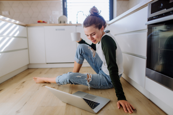 Young woman sitting with laptop and cup of coffee in her kitchen.