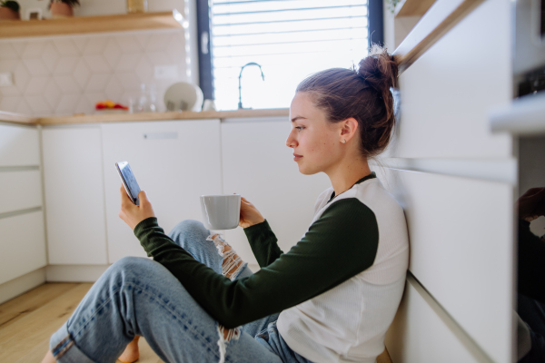 Young woman sitting with smartphone and cup of coffee in her kitchen.