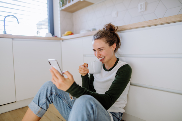 Young woman sitting with smartphone and cup of coffee in her kitchen.