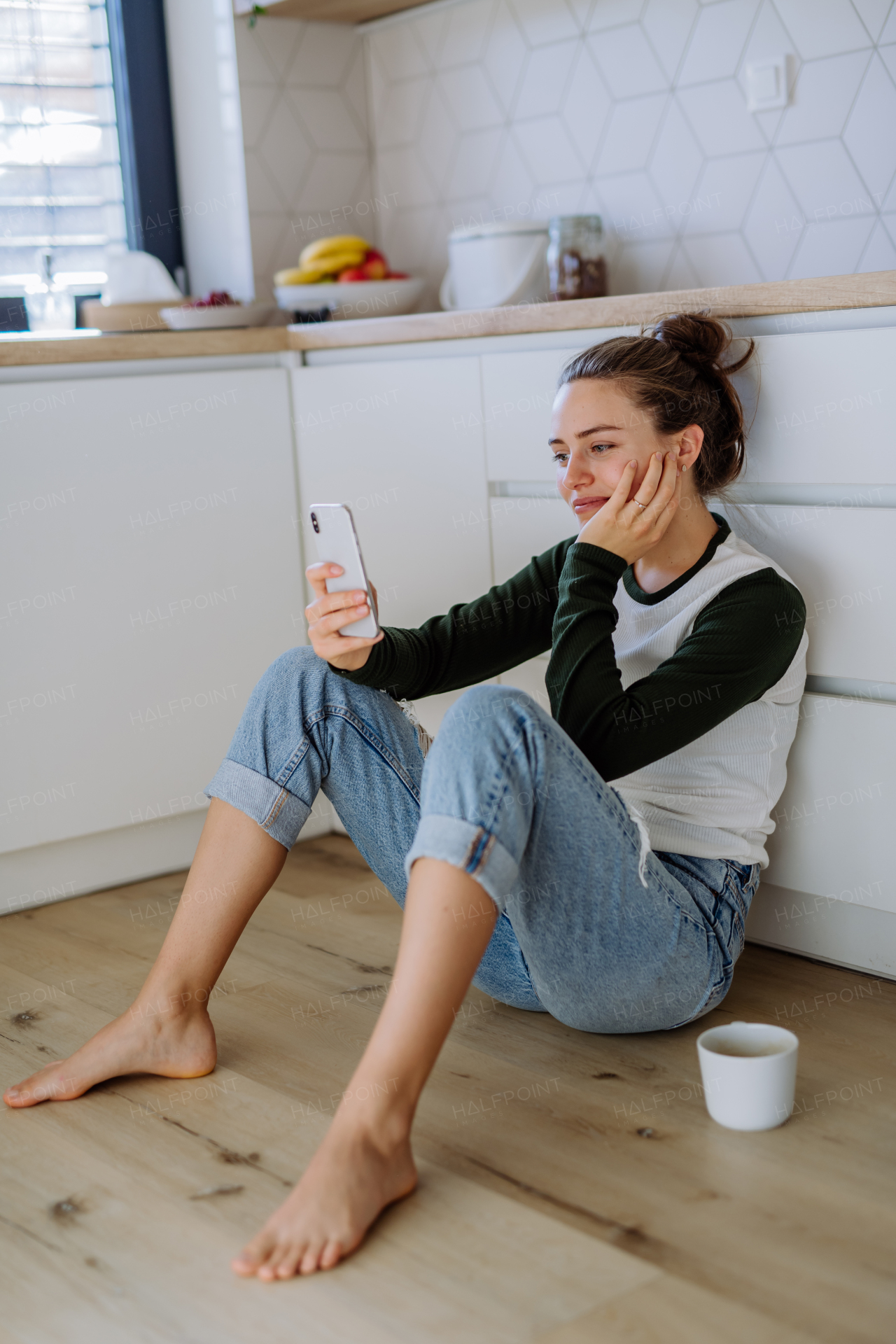 Young woman sitting with smartphone and cup of coffee in her kitchen.